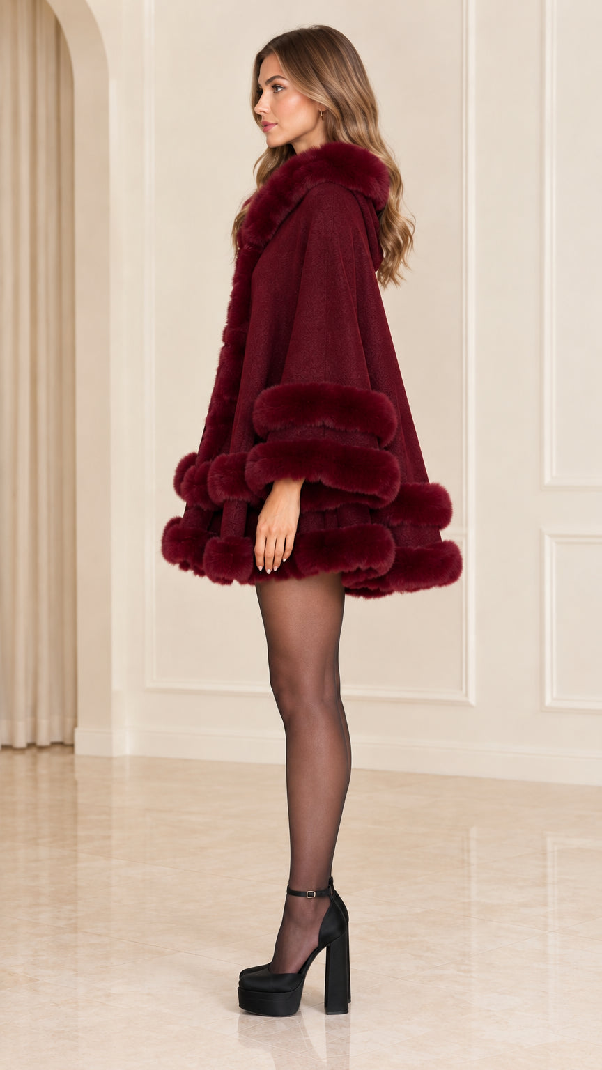 Side profile of woman wearing burgundy hooded cape coat with tiered fur hem