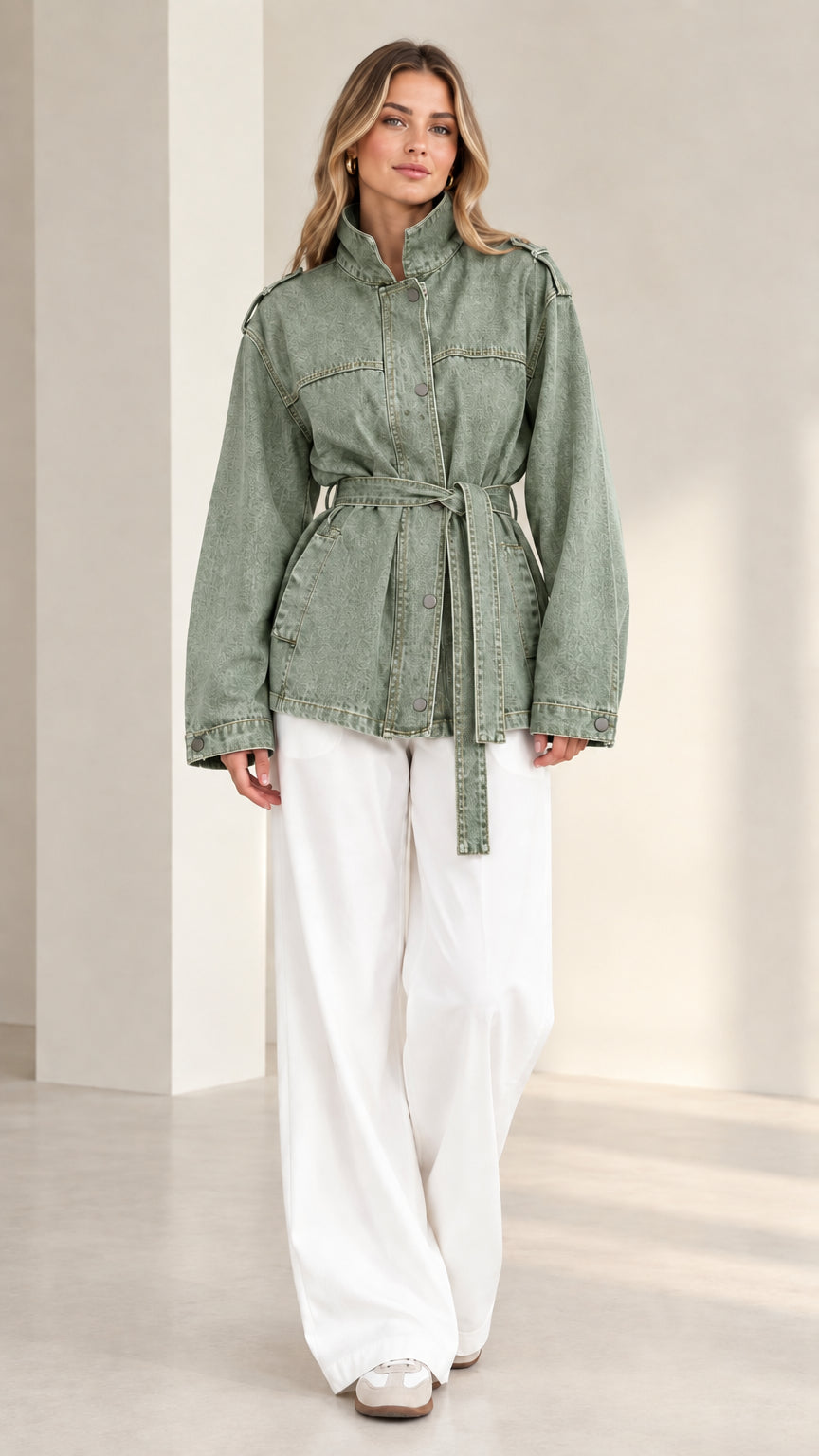 Front view of model wearing sage green belted safari denim jacket with white pants