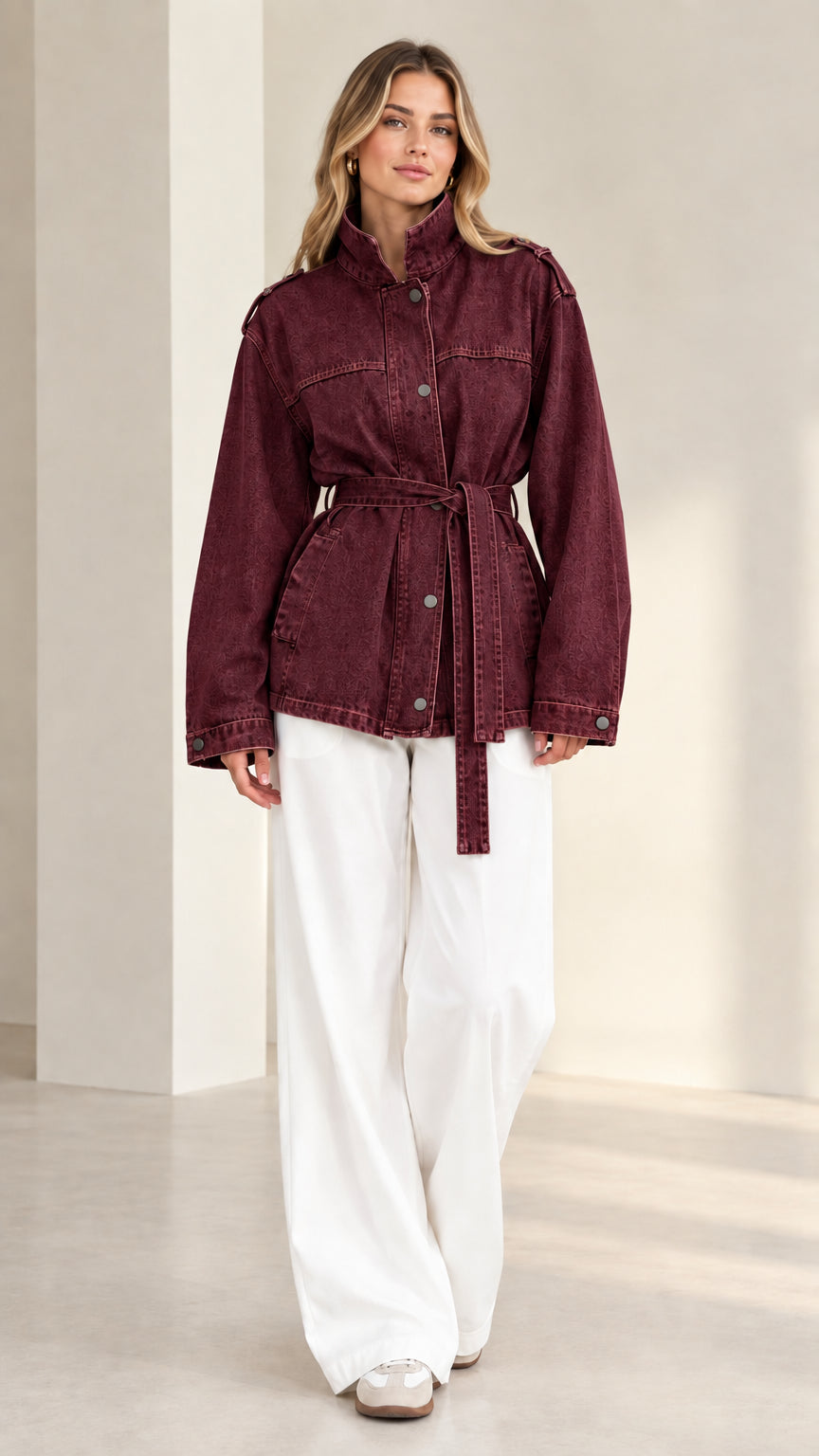 Front view of model wearing burgundy wine-washed belted denim jacket with white pants